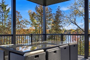 Un espace extérieur aménagé, abrité, avec une table à plateau de verre et des meubles en osier, offrant une vue pittoresque sur les arbres aux couleurs automnales et un plan d’eau.