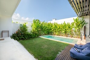 Ein moderner Poolbereich im Freien mit Holzterrasse, einer kleinen Rasenfläche und verschiedenen tropischen Pflanzen.