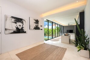 A modern living room with a large window, a kitchen area, and two abstract paintings on the wall. - Marea 2BR Luxury Private Pool Villa - Uluwatu (South Kuta)