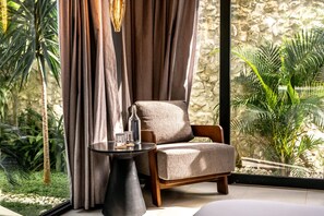 A cozy seating area with a cushioned chair, a small table with a bottle and glasses, and a view of tropical plants through a window with curtains. - Brezza 3BR Luxury Private Pool Villa - Uluwatu (South Kuta)