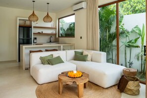 A modern living room with a white sofa, a wooden coffee table with fruit, and a kitchen area with a refrigerator and hanging lights. - Majo 1BR Romantic Villa, 3 Min to Thomas Beach (South Kuta)