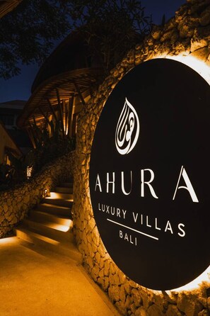 Naamplaatje voor 'AHURA LUXURY VILLAS' met een gestileerd logo, bij de ingang van een landgoed met stenen trappen en een overdekte doorgang.