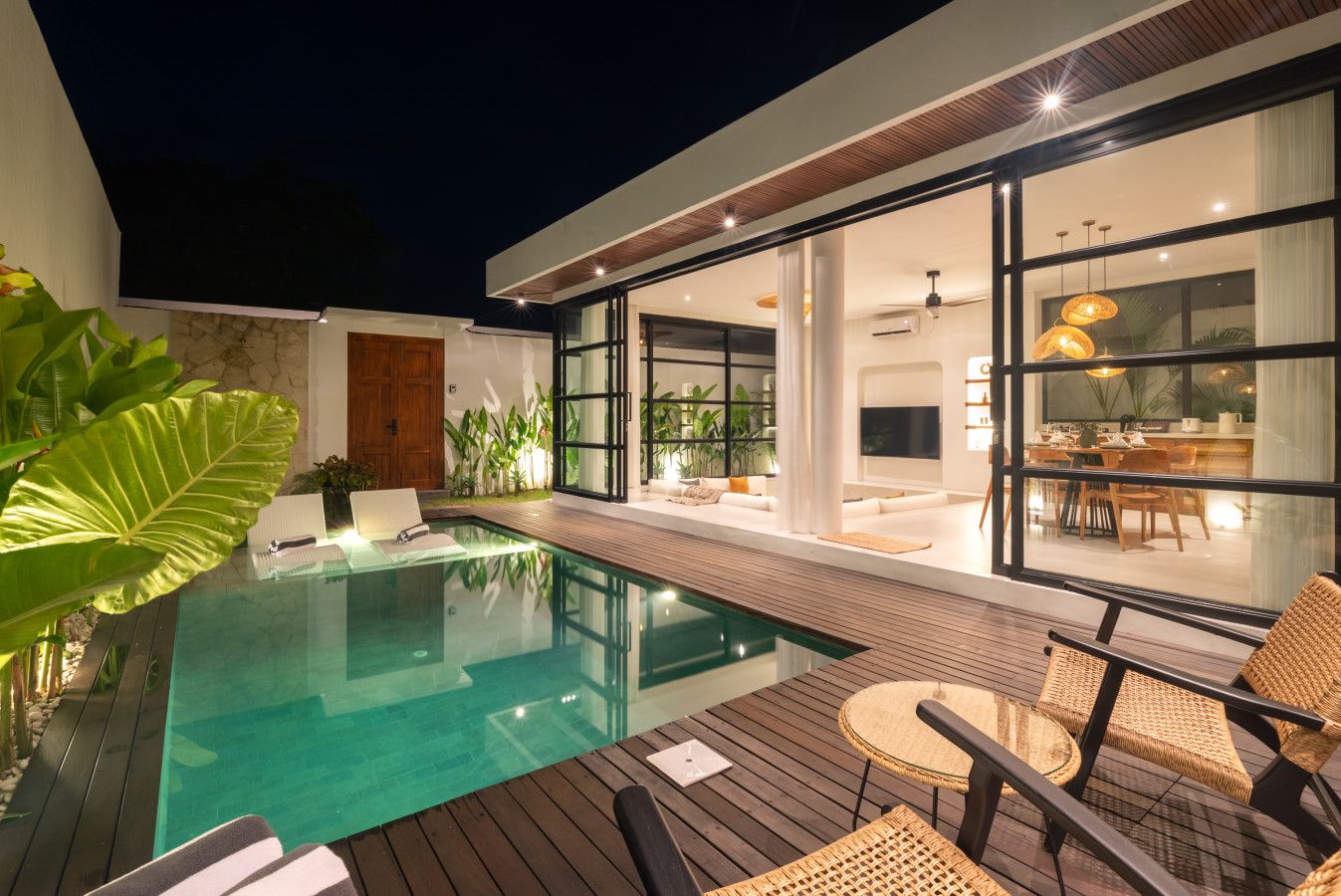 A modern outdoor pool area with a wooden deck, wicker furniture, and a glass-enclosed dining area.