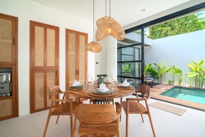 A modern dining area with a round wooden table set for four, surrounded by wooden chairs. There are two woven pendant lights above the table. The room features large glass doors leading to a pool area with tropical plants outside. - Villa Sofra 2BR Private Pool, 350mbps WiFi- Bingin (South Kuta)