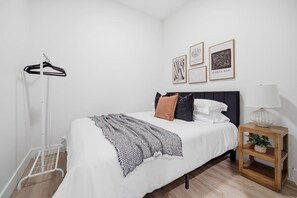 Ein ordentlich bezogenes Bett mit weißer Decke und einem gemusterten Überwurf, ein Nachttisch aus Holz mit einer Lampe und ein an der Wand befestigter Kleiderhaken.