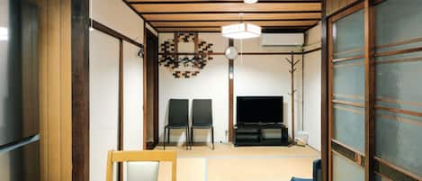 Quarto japonês tradicional com tatames, uma televisão e uma mesa de jantar com cadeiras.