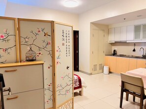 A room with a traditional sliding door, a bed with a red and white bedspread, a dining table with chairs, a kitchen area with cabinets and a sink, and a small trash bin. - One Uptown Residence BGC Modern BGC Home | 300Mbps + Workspace (Makati City)