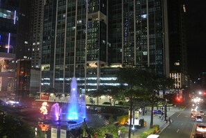 A nighttime cityscape with a large building featuring multiple floors and a fountain with illuminated water jets in the foreground. - One Uptown Residence BGC Modern BGC Home | 300Mbps + Workspace (Makati City)