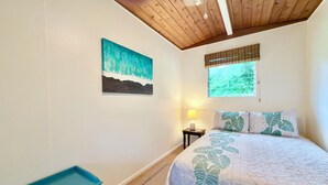 A bedroom with a bed, a nightstand, a lamp, a window with blinds, a painting on the wall, and a blue bench. - Private Gated Tropical Cottage Near Hilo & Pāhoa –Peaceful, Secure Island Escape (Keaau)