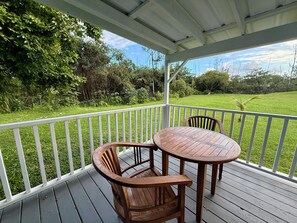 Área de estar coberta ao ar livre com mesa e cadeiras, cercada por um corrimão branco, com vista para uma paisagem gramada.