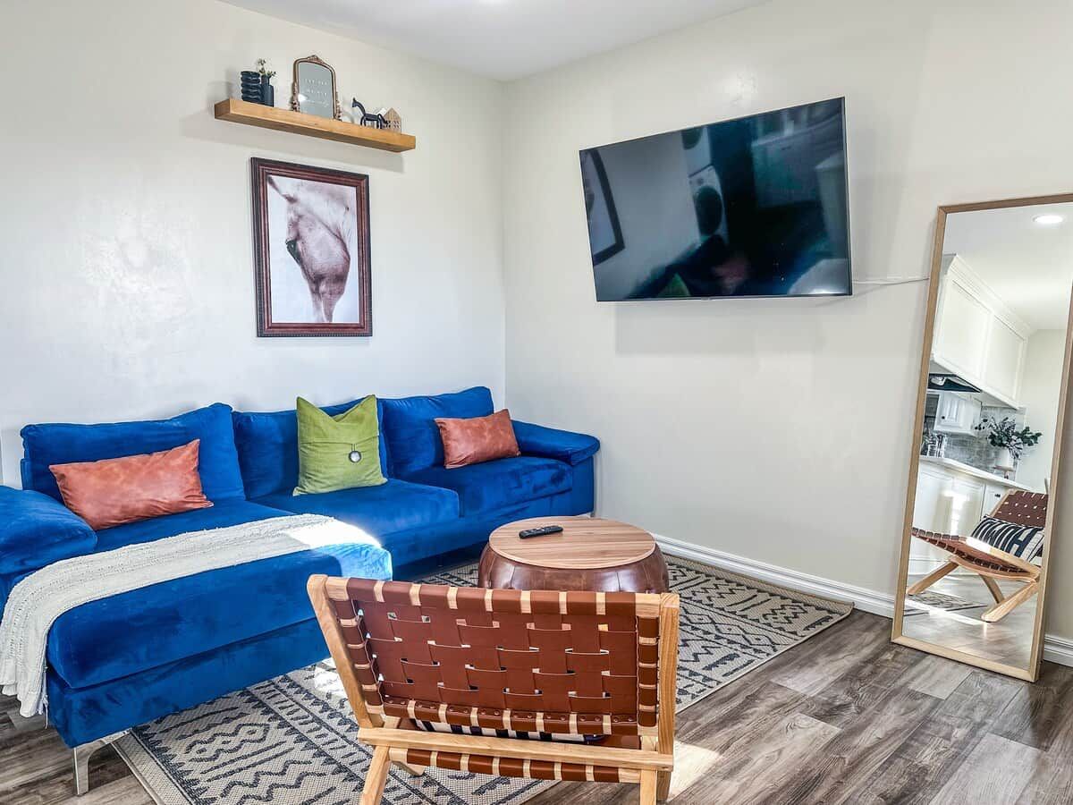 Un salon avec un canapé bleu, une chaise en bois, une table ronde en bois, un téléviseur fixé au mur et un miroir.