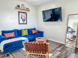 A living room with a blue sofa, a wooden chair, a round wooden table, a mounted TV, and a mirror.