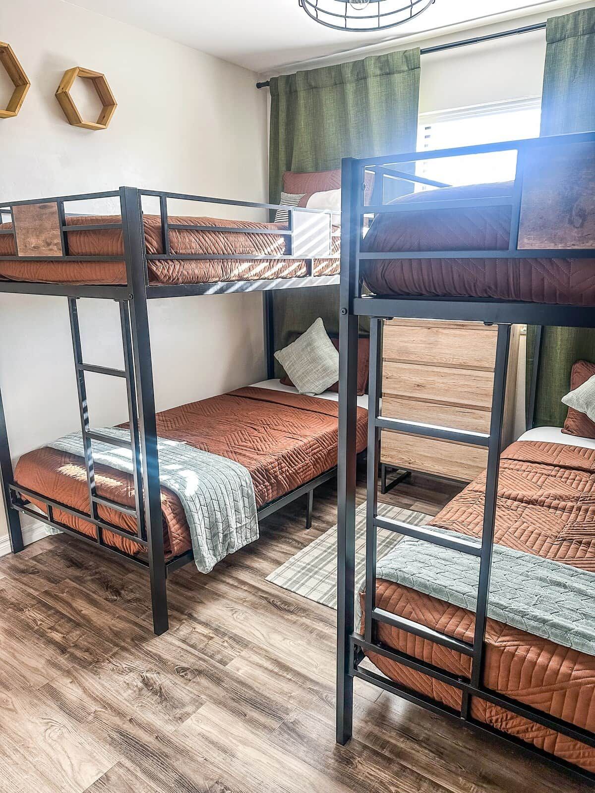Quarto com beliches, piso de madeira, janela com cortinas e decoração hexagonal na parede.