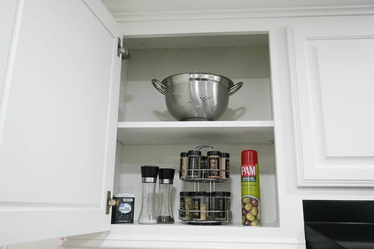 Un armario de cocina con un colador de acero inoxidable, frascos de especias y una lata de aceite en aerosol PAM.