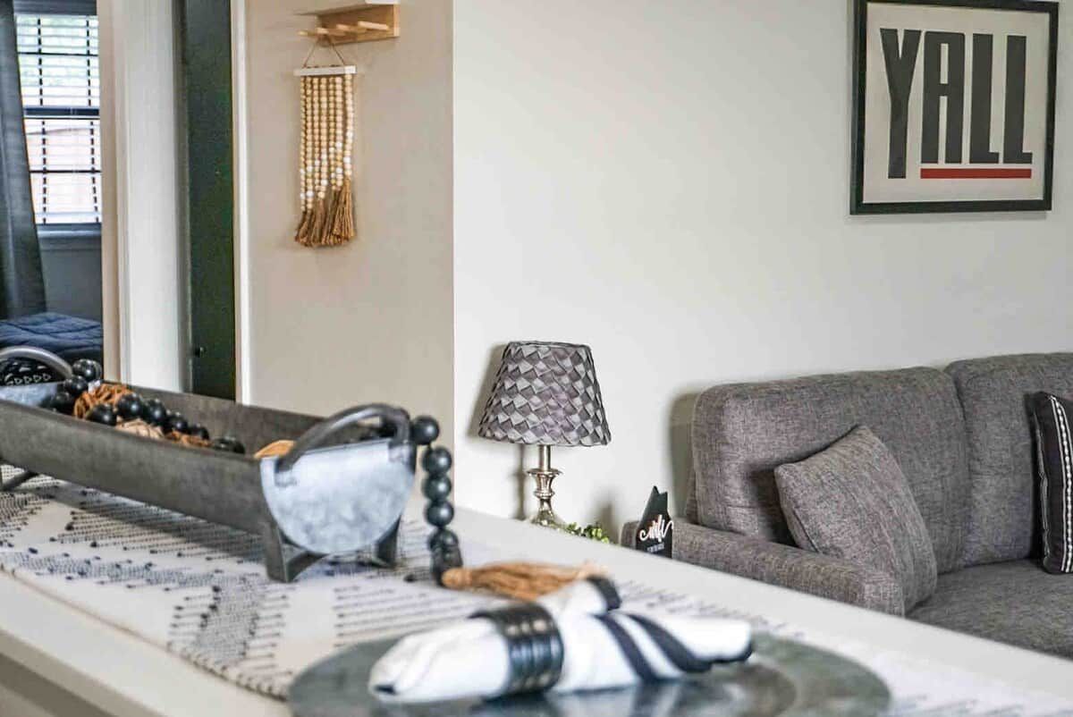 A living room with a grey sofa, a lamp, and a framed 'YALL' sign on the wall.