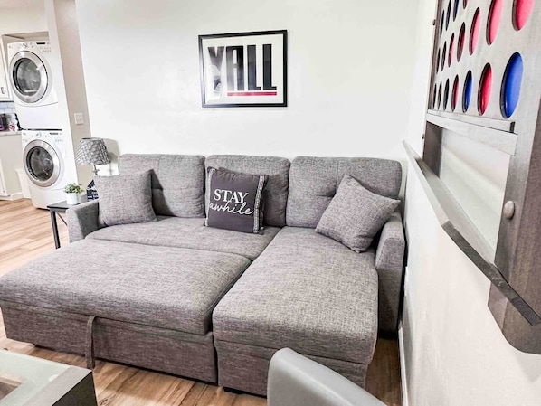 Un canapé d’angle gris, agrémenté d’un coussin sur lequel on peut lire : « Restez un peu ». La scène se déroule dans une chambre d’hôtel.