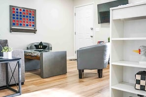 Un salon avec un baby-foot, un canapé gris, une porte blanche et une étagère sur laquelle se trouve un sac à carreaux.