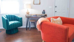 A room with a teal armchair, an orange sofa, a small table with books, a lamp, and a mirror on the wall. - Colorful Retreat | Near I-40 &SWOSU | Pets Welcome (Weatherford)