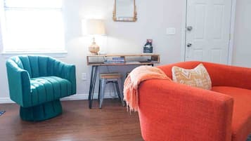Une pièce avec un fauteuil turquoise, un canapé orange, une petite table avec des livres, une lampe et un miroir au mur.