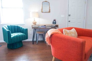 Une pièce avec un fauteuil turquoise, un canapé orange, une petite table avec des livres, une lampe et un miroir au mur.