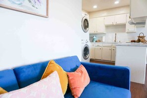 A living room with a blue sofa, a washing machine, and a kitchen area with white cabinets.