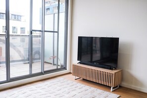 A room with a large window, a flat-screen TV on a wooden stand, and a white rug on the floor. - Stylish St Kilda Apartment | 2BR, 2 Bathrooms, Balcony + Secure Parking (St Kilda)