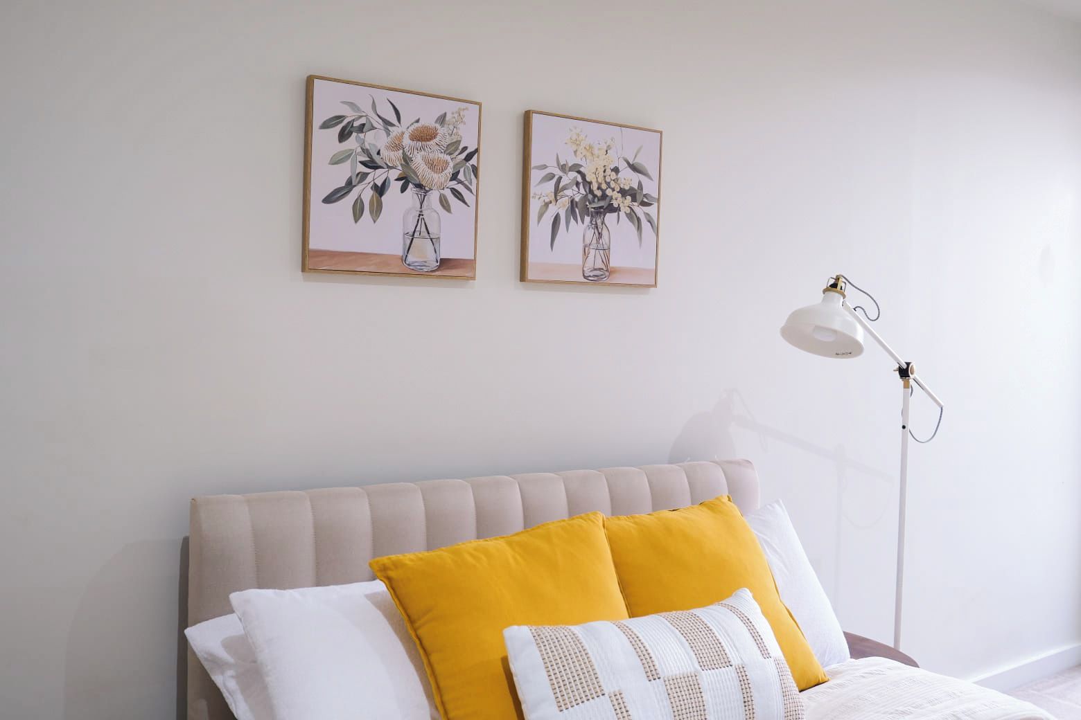A bedroom with a bed, two framed floral paintings, a bedside lamp, and a window.