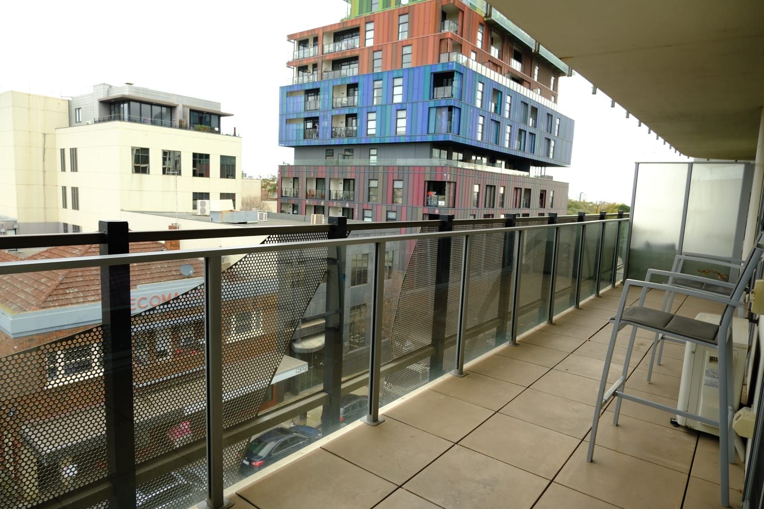 A balcony with a metal railing, a chair, and a view of multi-story buildings.