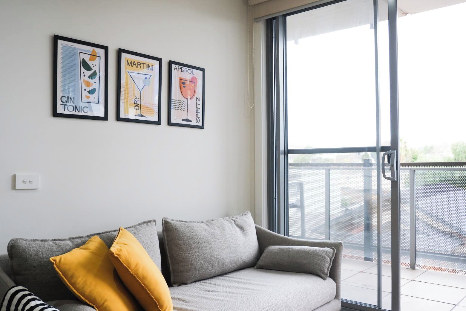 A modern living room with a grey sofa, a yellow pillow, and three framed wall art pieces depicting gin tonic, martini, and aperol.