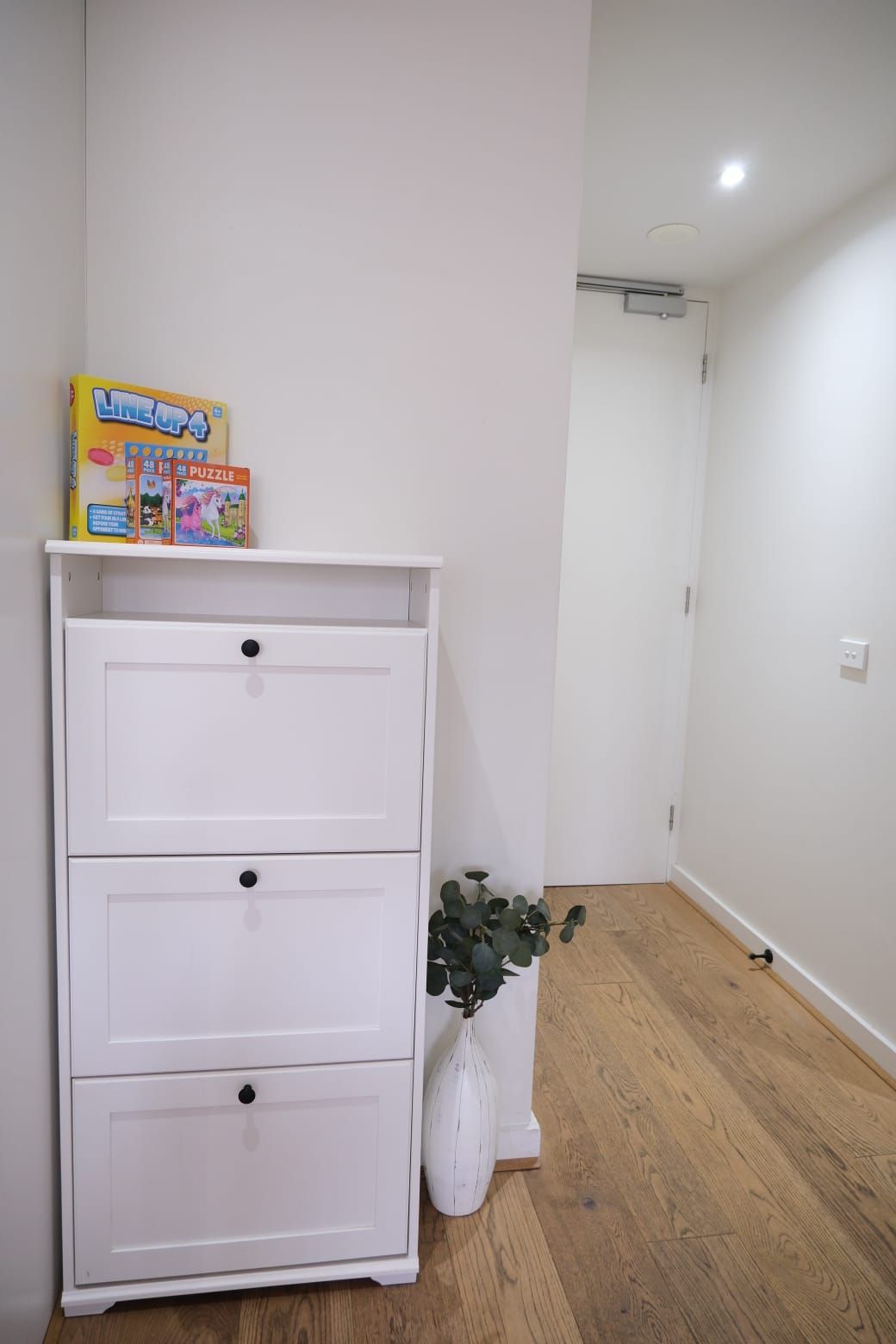 Une commode blanche, avec une plante placée à côté, dans une pièce au plancher en bois et aux murs blancs.