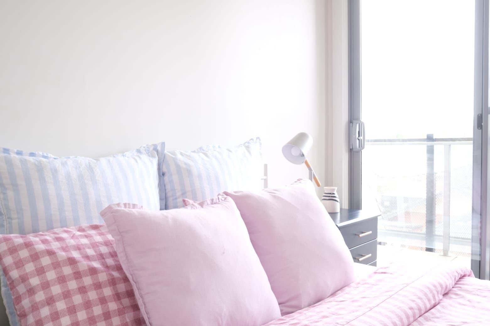 A bedroom with a bed, pillows, a nightstand, a lamp, and a view through a window.
