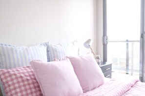 A bedroom with a bed, pillows, a nightstand, a lamp, and a view through a window. - Stylish St Kilda Apartment | 2BR, 2 Bathrooms, Balcony + Secure Parking (St Kilda)