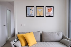 A neutral-toned living room with a grey sofa, two cushions, and three framed wall art pieces depicting gin, tonic, martini, and aperol. - Stylish St Kilda Apartment | 2BR, 2 Bathrooms, Balcony + Secure Parking (St Kilda)
