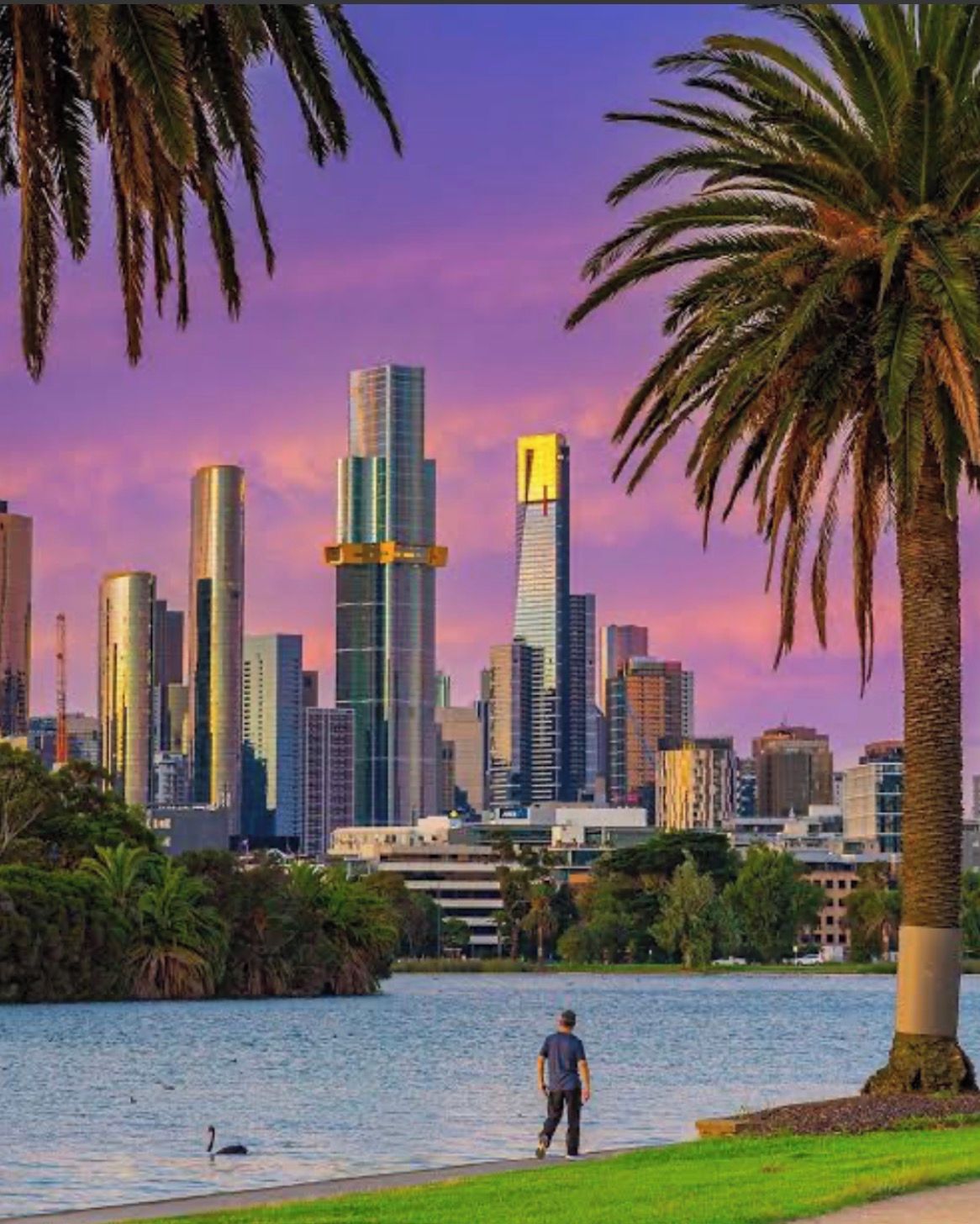A city skyline with modern skyscrapers, a body of water, and a person walking along the shore.