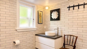Un baño con pared de ladrillo blanco, una ventana, un lavamanos blanco y una silla de madera.