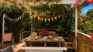 Una terraza exterior de madera con una mesa dispuesta para comer, rodeada de follaje y una cerca de madera.