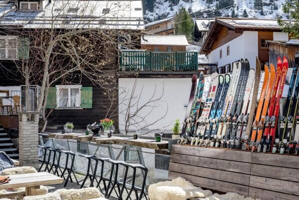 Une station de ski où l’on peut admirer une variété d’équipements de ski exposés à l’extérieur d’un bâtiment, ainsi que des espaces de repos en plein air et une vue sur les montagnes environnantes.