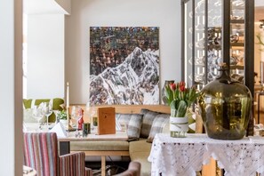 Un coin repas avec une table dressée pour deux personnes, un grand tableau représentant une montagne et une vitrine contenant des bouteilles.