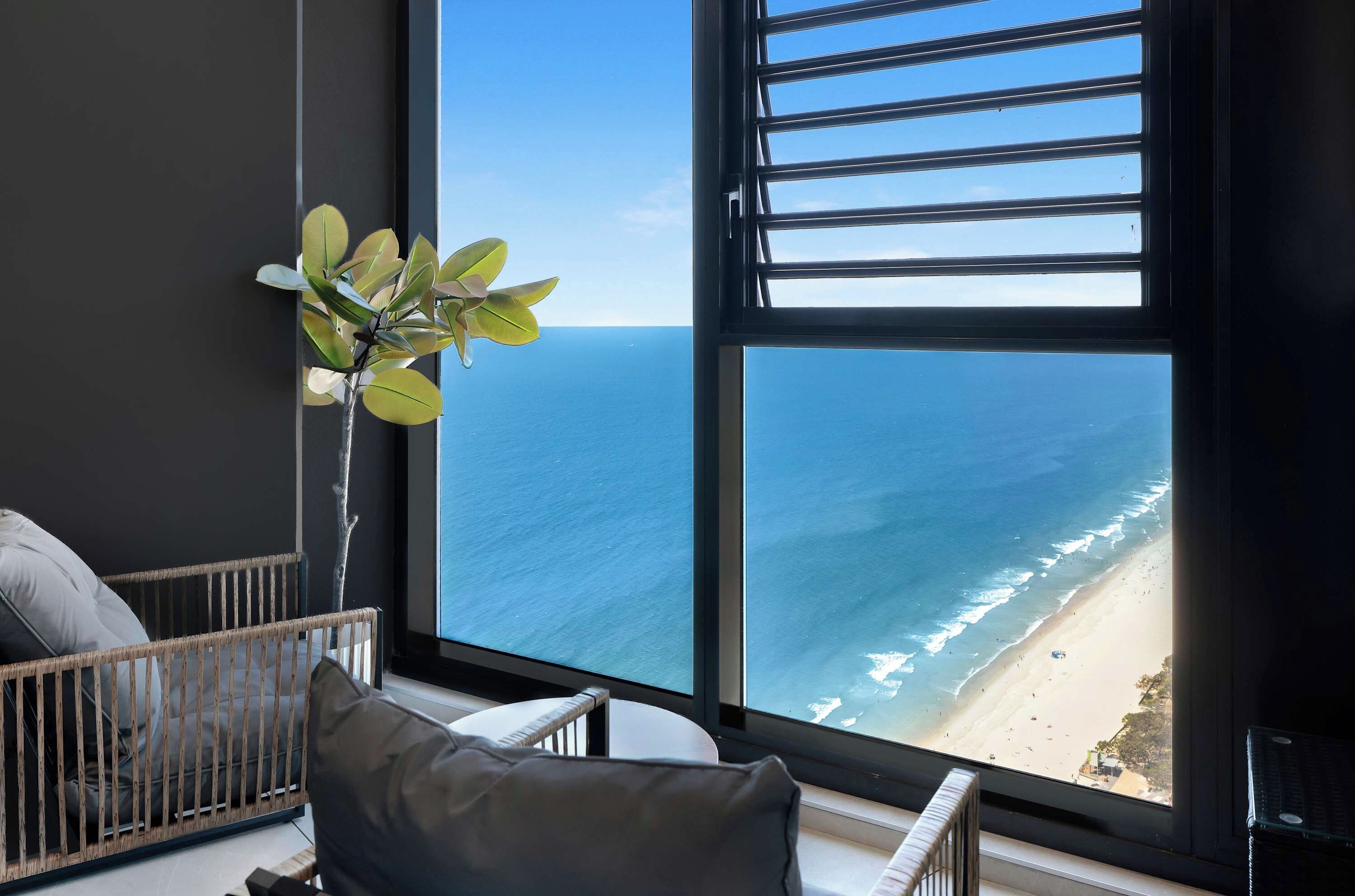 Ein Zimmer mit großem Fenster und Blick auf einen Strand, ein Korbstuhl und ein Blumentopf.
