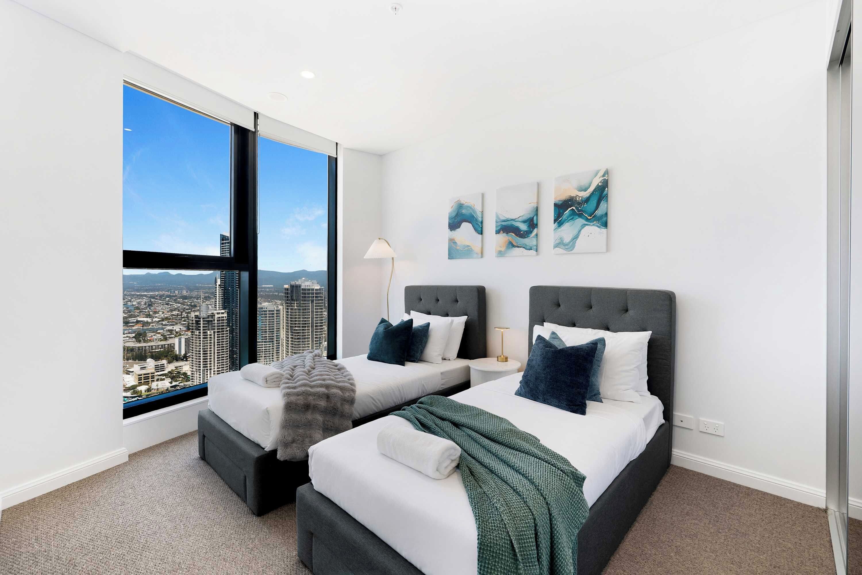 Une chambre d’hôtel moderne avec vue sur la ville, deux lits, une grande fenêtre et des œuvres d’art au mur.