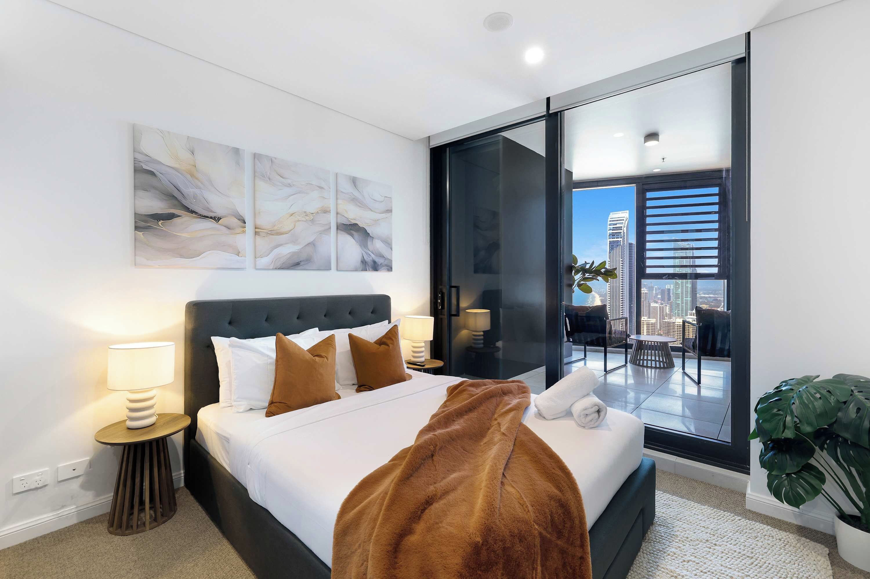 A modern hotel room with a large bed, a view of the city, and a balcony with a plant.