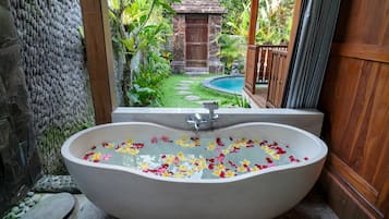 Eine freistehende Badewanne, gefüllt mit Wasser und Blütenblättern, vor einer Kulisse aus üppiger Bepflanzung und einer Steinmauer.