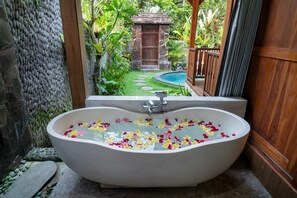 A freestanding bathtub filled with water and flower petals, set against a backdrop of lush greenery and a stone wall.
