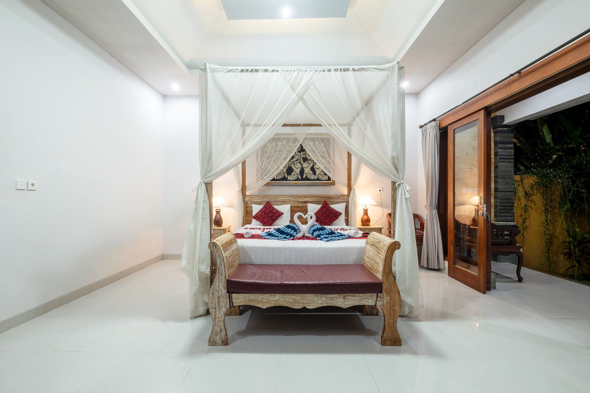 Una habitación con una cama bajo un dosel, un banco de madera, una lámpara de pared y un cuadro enmarcado.