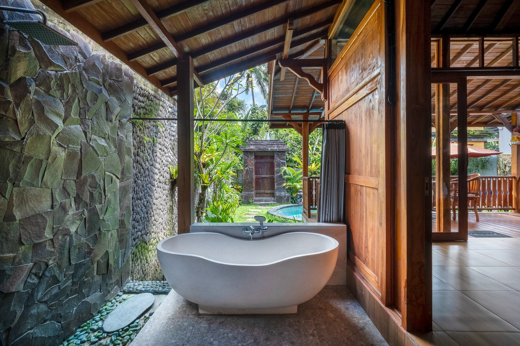 A freestanding bathtub in a covered outdoor area with stone walls and a view of a tropical garden.