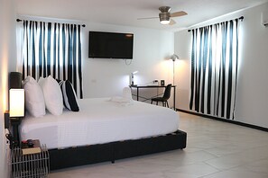 Une chambre d’hôtel avec un grand lit, un bureau, une chaise, un téléviseur fixé au mur et un ventilateur de plafond.