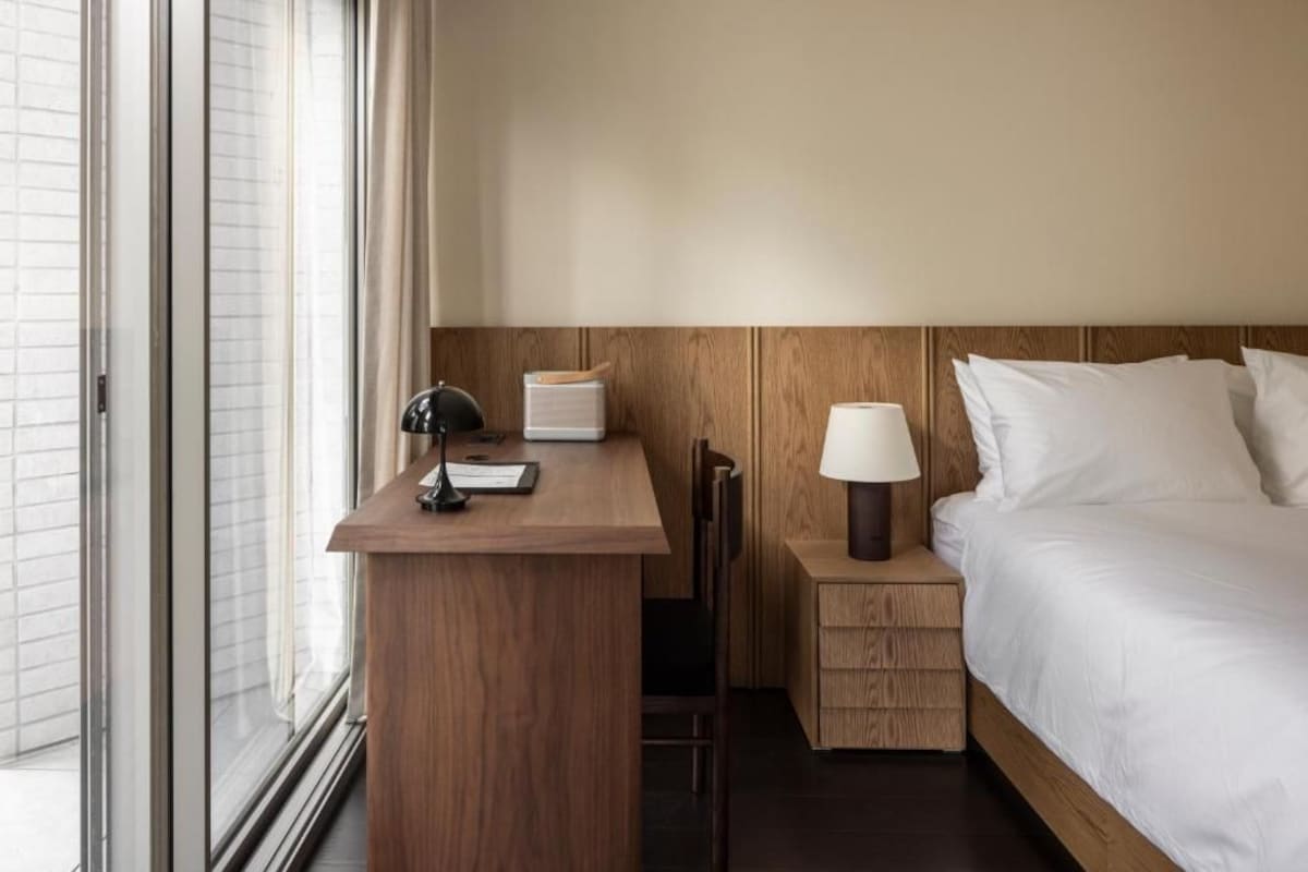 Quarto de hotel com escrivaninha de madeira, mesa de cabeceira com abajur, cama com roupa de cama branca e janela com persianas.