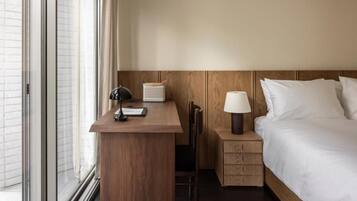 A hotel room with a wooden desk, a bedside table with a lamp, a bed with white linens, and a window with blinds.