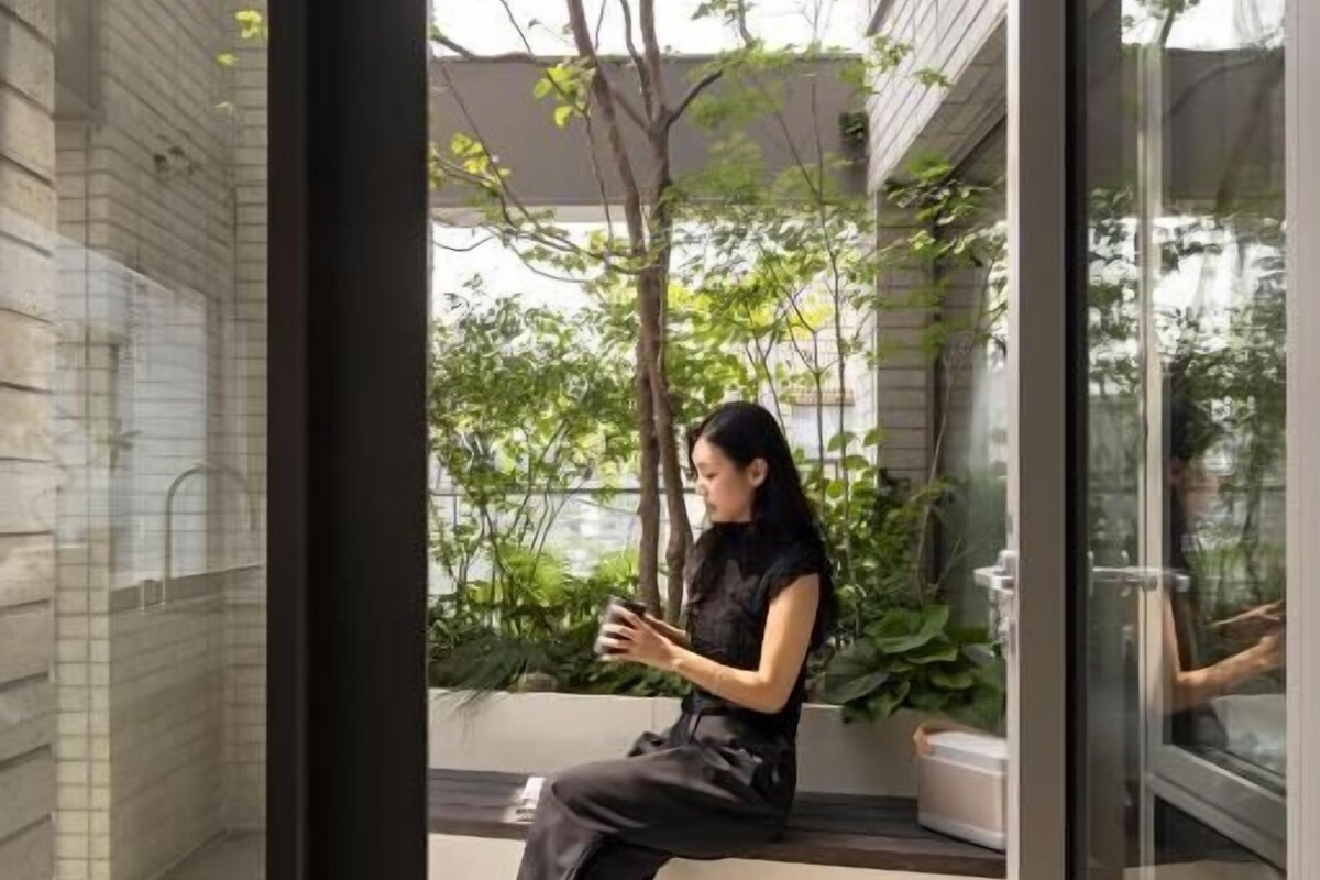 Una mujer sentada en un banco en un espacio interior moderno con paredes de vidrio, piso de baldosas y vistas al exterior con vegetación.