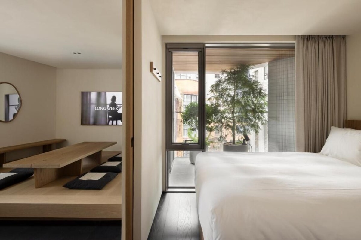 Quarto de hotel com uma cama, um banco, uma planta em vaso e vista para o exterior.